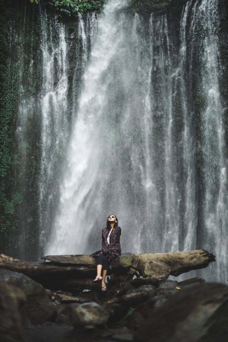 woman sitting fallen tree trunk in front of a waterfalls